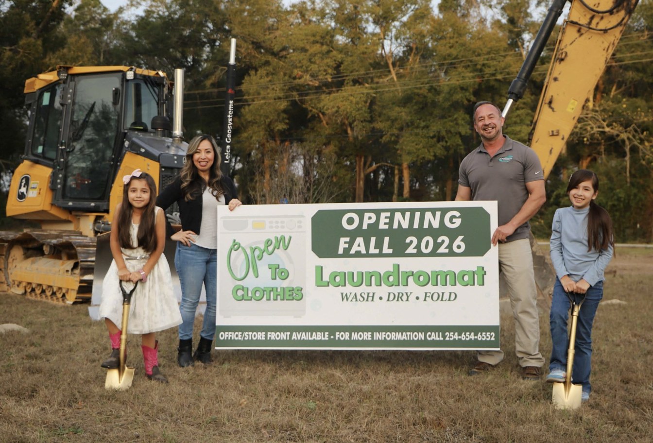 The owner's family at the construction site beside a sign reading Opening Fall 2026, Open To Clothes Laundromat, Wash Dry Fold